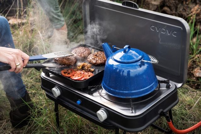 Person boiling water in a kettle and cooking burgers on a camping stove